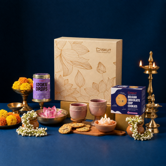 A beautifully arranged product display featuring a large beige BISKUT gift box with gold floral line art, alongside a can of Cookie Drops, a box of Belgian Chocolate Cookies, pink ceramic cups, cookies on a wooden board, marigold flowers, and brass oil lamps against a dark blue background.
