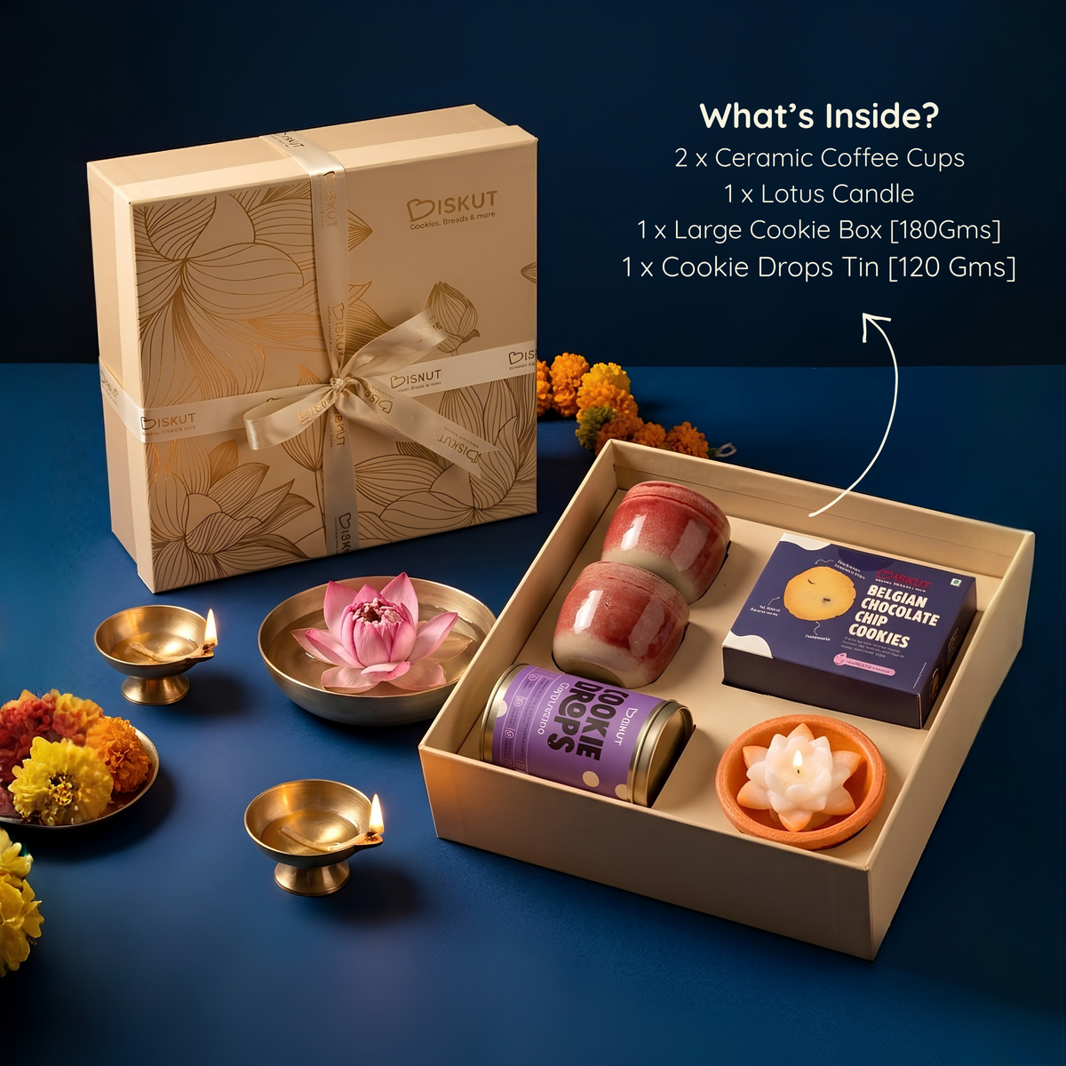 A gourmet gift set from BISKUT on a dark blue background, featuring a beige floral-patterned gift box, an open box with ceramic coffee cups, a purple chocolate tin, and a cookie tin, surrounded by marigold flowers and brass oil lamps. An 'What's Inside?' text overlay is visible on the top right.